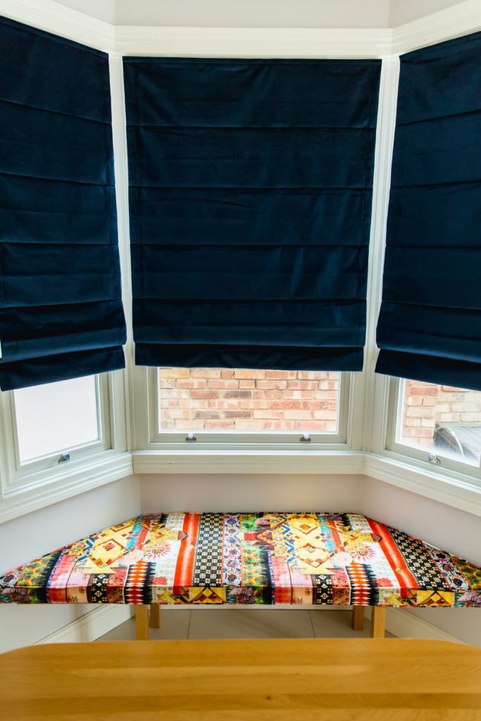 Blue roman blinds placed in a bay window above a colourful window bench